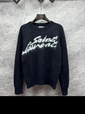 Svart Saint Laurent stickad tröja - Säljer en svart stickad tröja från Saint Laurent med vit logga i graffiti-stil på bröstet. Tröjan har rund halsringning, långa ärmar och ribbade muddar vid ärmslut och nederkant. Perfekt för dig som gillar streetwear och exklusiva märken.