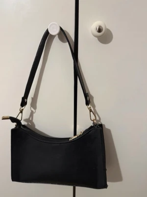 Svart handväska med gulddetaljer - Stilren svart handväska i avlång modell med guldfärgade metalldetaljer och dragkedja. Väskan har ett avtagbart axelband och minimalistisk design som passar till många olika looks.