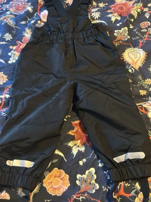 Svarta täckbyxor från Kaxs Proxtec - Svarta täckbyxor från Kaxs Proxtec med justerbara hängslen och resår i midjan. Byxorna har reflexdetaljer vid bensluten och är tillverkade i slitstarkt material, perfekta för kalla dagar utomhus.