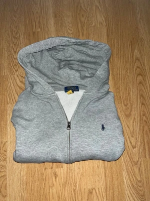 Grå zip hoodie från Polo Ralph Lauren - Snygg grå hoodie med dragkedja från Polo Ralph Lauren. Klassisk design med huva och det ikoniska lilla blå logobroderiet på bröstet. Tillverkad i mjukt bomullsmaterial som är skönt att ha på sig. Perfekt för en avslappnad och stilren look. Bara hör av er vid funderingar!😃