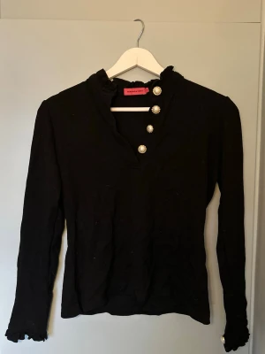 Svart blus med pärlknappar Veronica Virta - Svart blus/topp från Veronica Virta med lång ärm och dekorativa pärlknappar. Blusen har volangdetaljer vid kragen och är tillverkad i ett ganska tungt mjukt tyg.