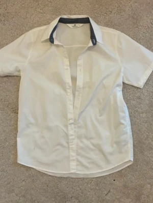 Vit kortärmad skjorta med kontrastkrage - Klassisk vit skjorta i bomull med korta ärmar och knappar framtill. Skjortan har en snygg kontrastfärgad krage i mörkblått som ger en modern touch. Perfekt till jeans eller chinos för en clean look.