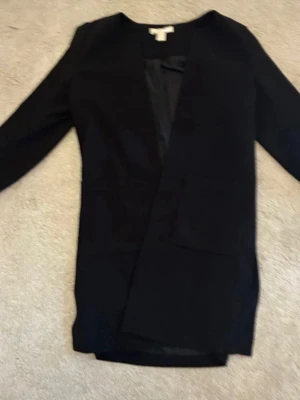 Svart kavaj med öppen front - Stilren svart kavaj med öppen front och lång ärm. Klassisk rak passform utan synliga knappar eller dragkedja. Perfekt att slänga över en t-shirt eller skjorta för en clean look. Materialet känns mjukt och följsamt.