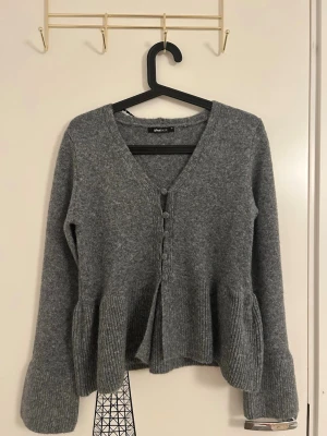 Grå kofta med volang från Gina Tricot  - Grå kofta med volangkant nedtill och vid ärmslut från Gina Tricot i storlek XS. Koftan har v-ringning och knappar framtill. Tillverkad i mjukt stickat material. Aldrig använd men har inte prislappen kvar.