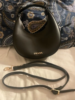 Svart handväska  - Snygg svart handväska från P R A D A i slätt fejk skinn med guldfärgade detaljer och avtagbar axelrem. Väskan har en rundad form och diskret P R A D A-logga framtill. Perfekt accessoar för att lyfta din outfit. OBS! Ej äkta köpt i Spanien för 50€