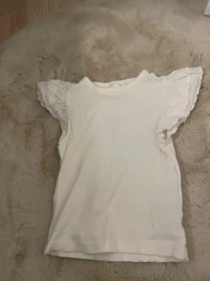 Vit topp med volangärmar - Söt vit topp med ribbad struktur och korta volangärmar i spets. Perfekt för dig som gillar romantiska detaljer och en ljus, fräsch look. Toppen har rund halsringning och är tillverkad i ett mjukt material som känns skönt mot huden.