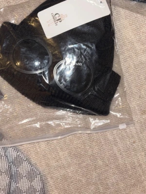 Svart mössa med glasögondetalj - Säljer en svart ribbstickad mössa från C.P. Company med ikoniska glasögonlinser framtill. Mössan är tillverkad i mjukt material och har en modern, streetinspirerad look som sticker ut.