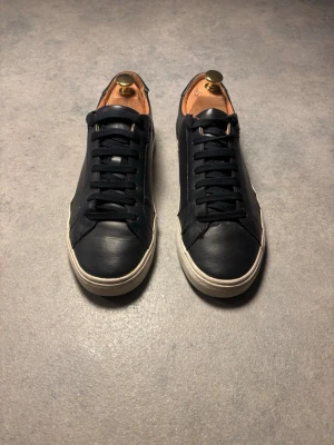 Svarta/Mörkblå sneakers från Zara - Stilrena svarta/mörkblå sneakers i skinn med klassisk rund tå och vit platt sula. Skorna har svarta snören och diskreta detaljer, perfekta för en clean look.