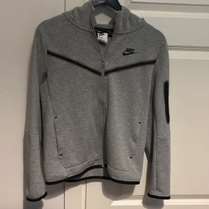 Grå Nike tech hoodie med dragkedja - Snygg grå hoodie från Nike med hel dragkedja och svart Nike-logga på bröstet. Tröjan har huva, svarta detaljer, långa ärmar och praktisk ficka med dragkedja på ena ärmen. Perfekt för chill eller träning. Nypris ca 900kr 