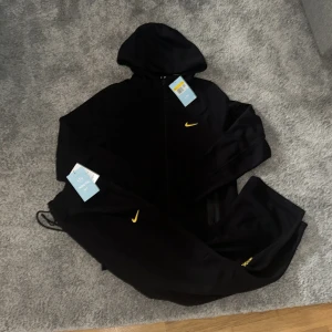 Svart NOCTA Nike vindjacka med huva - Svart vindjacka från Nike NOCTA med huva och hel dragkedja. Jackan har en gul Nike-logga på bröstet och är tillverkad i polyester. Snygg och sportig design med loose passform, perfekt för dig som gillar streetwear och vill sticka ut med detaljer i gult.