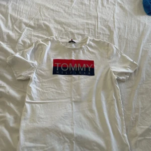 Vit Tommy Hilfiger t-shirt med tryck - Snygg vit t-shirt från Tommy Hilfiger med stort logotryck i rött och blått framtill och vertikalt tryck på ryggen. Klassisk passform och rund hals. Perfekt för dig som gillar stilrena och ikoniska plagg.