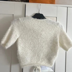 Vit fluffig stickad tröja med knyt - Supermjuk och fluffig vit stickad tröja från na-kd med korta ärmar och knytband baktill. Tröjan har en croppad passform och är perfekt för dig som gillar en söt och trendig look. Materialet är stickat och känns riktigt cozy.