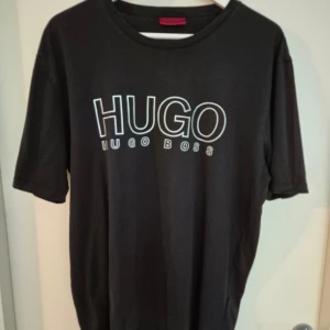 Svart t-shirt från Hugo Boss - Svart t-shirt från Hugo Boss med stor vit HUGO-logga framtill. Klassisk rund halsringning och korta ärmar. Skön passform och tillverkad i mjuk bomull, perfekt för en clean och stilren look.