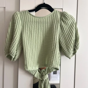 Grön omlottopp med puffärmar - Säljer en ljusgrön omlottopp med puffiga korta ärmar och knyt i baktill. Toppen har en strukturerad, våfflad yta och är croppad. 