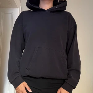 Svart hoodie med huva och ficka - En klassisk hoodie med huva och en stor magficka. Tröjan har långa ärmar. Den är något sliten i svart färg på grund av användning.