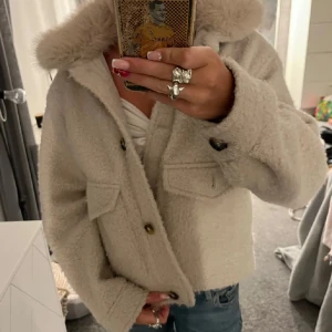 Beige teddyjacka med fuskpäls - Supermysig beige teddyjacka med fluffig fuskpäls runt kragen. Jackan har stora fickor framtill, knappar och en lite boxig, croppad passform. Perfekt för kalla dagar när du vill vara både varm och snygg.