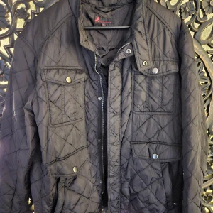 Svart quiltad jacka Dressmann XL - Svart quiltad jacka från Dressmann i storlek XL. Jackan har flera fickor med tryckknappar, dragkedja framtill och en klassisk krage. Snyggt rutmönster i tyget och en stilren look som passar till många outfits.