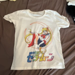 Vit Sailor Moon t-shirt  - Vit t-shirt med färgglatt Sailor Moon-tryck framtill och japansk text under motivet. Klassisk rund hals och korta ärmar. Perfekt för dig som älskar anime och vill sticka ut med en ikonisk look. Köpt i Japan