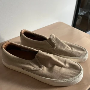 Beige slip-on sneakers från Stockholm Design Group - Stilrena slip-on sneakers i beiget från Stockholm Design Group. Skorna har vita sulor, detaljer i ljust skinn vid häl och insida, samt en enkel och clean design som passar till det mesta. Använda fåtal gånger så fortfarande i mycket bra skick.