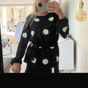 Svart prickig blus från Vila - Säljer en svart blus från Vila med stora vita prickar. Blusen har lång ärm, rund halsringning och ett knytband i midjan som ger en snygg siluett. Perfekt för dig som gillar statement-mönster och vill sticka ut lite extra.