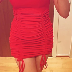 Röd tight kortklänning med snörning - Snygg röd kortklänning med tight passform och coola snörningsdetaljer på sidorna. Klänningen har korta ärmar och rynkade partier längs hela kroppen som ger en riktigt trendig look. Perfekt för dig som vill sticka ut med en färgstark och figurnära stil.