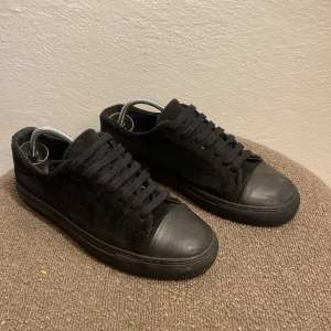 Svarta Arigato mockasneakers med gummitå - Snygga helsvarta sneakers från Arigato i mocka med tå och sula i gummi. Klassisk låg modell med rund tå och svarta skosnören. Perfekta för dig som gillar stilrena och minimalistiska skor.