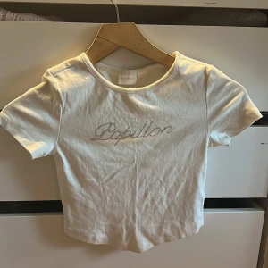 Vit t-shirt - Snygg vit t-shirt från Gina Tricot med korta ärmar. Framsidan har texten 'Papillon' i glittriga stenar som ger en extra touch. Perfekt till jeans eller kjol för en trendig look.