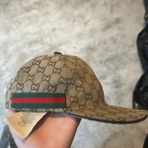 Gucci beige keps med GG-mönster - Snygg beige keps från Gucci med klassiskt GG-monogram över hela kepsen. Har gröna och röda ränder på sidorna och justerbar rem baktill. Materialet är bomull och insidan är mörkbrun. Perfekt för dig som vill ha en lyxig och trendig accessoar.