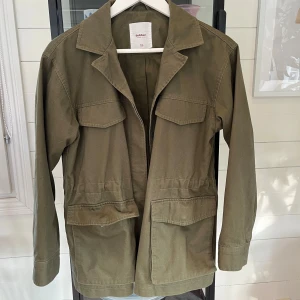 Olivgrön parkajacka från Dobber XS - Säljer en olivgrön parkajacka från Dobber i storlek XS. Jackan har fyra stora fickor framtill, klassisk krage och snörning i midjan för att justera passformen. Tillverkad i bomull och har långa ärmar. Perfekt för dig som gillar utility-vibe.