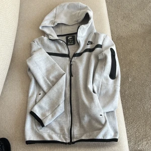 Grå hoodie från Nike med dragkedja - Säljer en grå hoodie från Nike med svart dragkedja och svarta detaljer. Tröjan har huva, lång ärm och en praktisk  med dragkedja på ena ärmen. Perfekt för dig som gillar sportig och avslappnad stil.