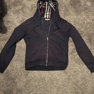 Mörkblå Burberry hoodie med dragkedja - Snygg mörkblå hoodie från Burberry med klassiskt rutigt foder i huvan och broderad logga på bröstet. Tröjan har dragkedja, snörning i huvan och känguruficka. Perfekt för dig som vill ha en stilren och lyxig look.