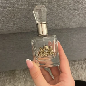 Juicy Couture parfymflaska - Snygg parfym från Juicy Couture i en genomskinlig fyrkantig glasflaska med silverfärgad hals och en stor, kristalliknande kork. Flaskan har en guldig emblem med krona och lejon på framsidan. Perfekt som statement på sminkbordet. Lite kvar
