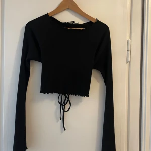 Svart croppad långärmad topp med snörning, NAKD - Säljer en svart croppad topp med långa ärmar som är vida och ribbad struktur. Toppen har öppen rygg med snygg snörning och volangkant vid ärmslut och nederkant. Det är storlek S men den är stretchig och passar därför bra för större storlekar också! Perfekt för dig som gillar trendiga detaljer!!