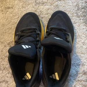 Svarta Adidas sneakers med gula detaljer - Säljer ett par snygga Adidas sneakers i svart med gula och rosa detaljer på sulan. Skorna har snörning och är tillverkade i syntetmaterial med ventilerande mesh upptill. Perfekta för dig som gillar sportig stil och vill sticka ut lite extra.