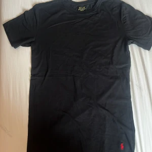 Svart t-shirt från Polo Ralph Lauren - Svart t-shirt från Polo Ralph Lauren med klassisk röd broderad logga vid nederkanten. Modellen är slim fit och har korta ärmar. Tillverkad i mjuk bomull som känns skön mot huden. Perfekt för dig som gillar stilrena och tidlösa plagg.