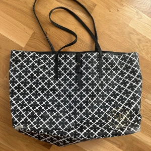 Svartvit shoppingväska By Malene Birger - Snygg shoppingväska från By Malene Birger i svart och vitt med klassiskt grafiskt mönster och guldfärgad logga framtill. Väskan har långa svarta handtag, innerficka med dragkedja och är tillverkad i tåligt syntetmaterial. Sliten i hörnen och fläckig på insidan som säkert går att tvätta bort. Säljs i befintligt skick.