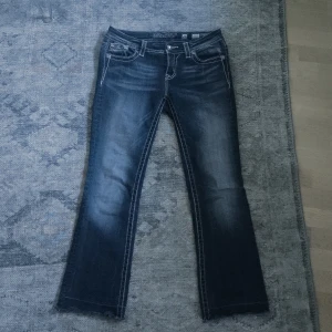 Miss Me bootcut lågmidjade jeans - Snygga blå lågmidjade jeans från Miss Me i bootcut-modell med broderade och glittriga detaljer på bakfickorna. Tydliga kontrastsömmar, byxorna är lite slitna och avklippta där nere och jeansen är i storlek w26.