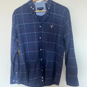 Blårutig skjorta från GANT, storlek L - Snygg blårutig skjorta från GANT med klassisk button-down krage och broderad logga på bröstet. Skjortan har vita knappar och långa ärmar. Perfekt för dig som gillar en stilren och tidlös look.