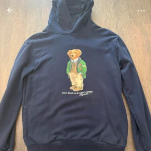 Marinblå hoodie Polo Bear Ralph Lauren - Snygg marinblå hoodie från Ralph Lauren med ikoniska Polo Bear-trycket på bröstet. Tröjan har huva, långa ärmar och mjukt material som känns skönt mot huden. Perfekt för dig som gillar klassisk streetstyle med en twist.