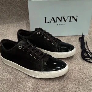  Lanvin skor - Snygga svarta sneakers från Lanvin med vit sula och glansig lackad tå. Skorna har snörning, metallöljetter och en mix av mocka och skinn. Perfekta för dig som gillar stilrena men ändå unika detaljer. Extra skosnören medföljer.