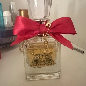 Juicy Couture parfym  - Juicy Couture parfym i en lyxig fyrkantig glasflaska med gulddetaljer och emblem framtill. Flaskan har en stor, glittrig kristallkork och är dekorerad med en röd sidenrosett och ett guldigt J-hänge. Doften är klassiskt feminin och flaskan är riktigt snygg på hyllan. 50 ml