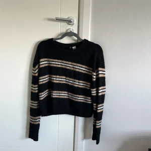 Svart randig stickad tröja - Mysig svart stickad tröja med beige och vita horisontella ränder. Tröjan har rund halsringning och långa ärmar. Perfekt för kyliga dagar och enkel att matcha med jeans eller kjol.