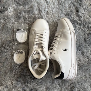 Vita sneakers Polo Ralph Lauren - Snygga vita sneakers från Polo Ralph Lauren i skinn med svart detalj på hälen och klassisk logga på sidan. Skorna har rund tå, platt sula och snörning. Medföljer två extra par vita skosnören. Perfekta för en clean och stilren look. Använda ett fåtal gånger bara.