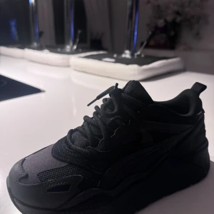 Svarta chunky sneakers från Puma - Säljer ett par svarta chunky sneakers från Puma med RS Running System. Skorna har en cool, sportig vibe med mesh och syntetdetaljer, samt kraftig sula och snörning. Perfekta för dig som gillar streetstyle och vill ha en bekväm och trendig look.