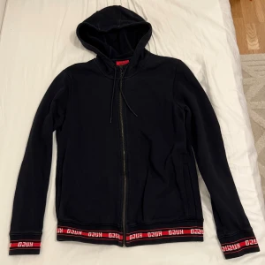 Säljer Hugo Boss Hoodie! - Svart hoodie från Hugo med hel dragkedja framtill och huva med snörning. Ribbade muddar och nederkant med röd och vit Hugo-logga. Storlek: XS 