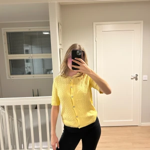 Gul kabelstickad kofta med kort ärm - Superfin gul kabelstickad kofta med korta ärmar och rund hals. Koftan har snygga knappar framtill och ribbade muddar vid hals och nederkant. Perfekt för dig som vill ha en färgklick i garderoben och gillar klassisk stil med en twist. Prislappen är inte kvar men tröjan har alldrig använts
