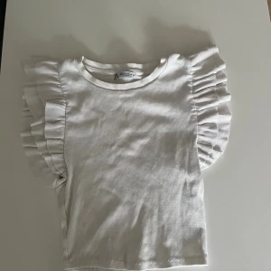 Vit volangtopp från Zara - Supersöt vit topp från Zara med ribbad struktur och stora volangdetaljer på axlarna. Perfekt för dig som vill ha något extra till outfiten! T-shirten är ärmlös och har en rund halsringning.