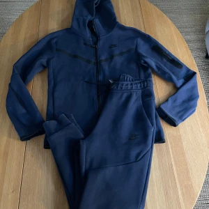 Nike Tech sätt - Marinblå . Jacka str S , Byxa str XS  - Nike Tech sätt , hoodiejacka o byxa   hel dragkedja och huva. Jackan har svarta detaljer, diskret Nike-logga på bröstet och en extra ficka med dragkedja på ärmen. Tillverkad i mjukt material, perfekt för chill eller träning. Säljas som sätt . 