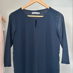 Mörkblå blus från Fransa - En stilren mörkblå klänning från Fransa med trekvartsärm och rund halsringning. Blusen har en liten knappdetalj vid halsen och är tillverkad i ett lätt, semitransparent material som ger en luftig känsla. Perfekt för dig som gillar enkel och klassisk design.