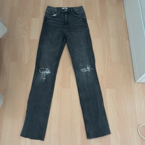 Grå jeans med slits från Zara - Snygga grå jeans från Zara i rak modell med slitna detaljer på knäna och slits nertill på benen. Jeansen har klassisk femficksdesign och är tillverkade i denim. Perfekta för dig som gillar en lite edgy stil.